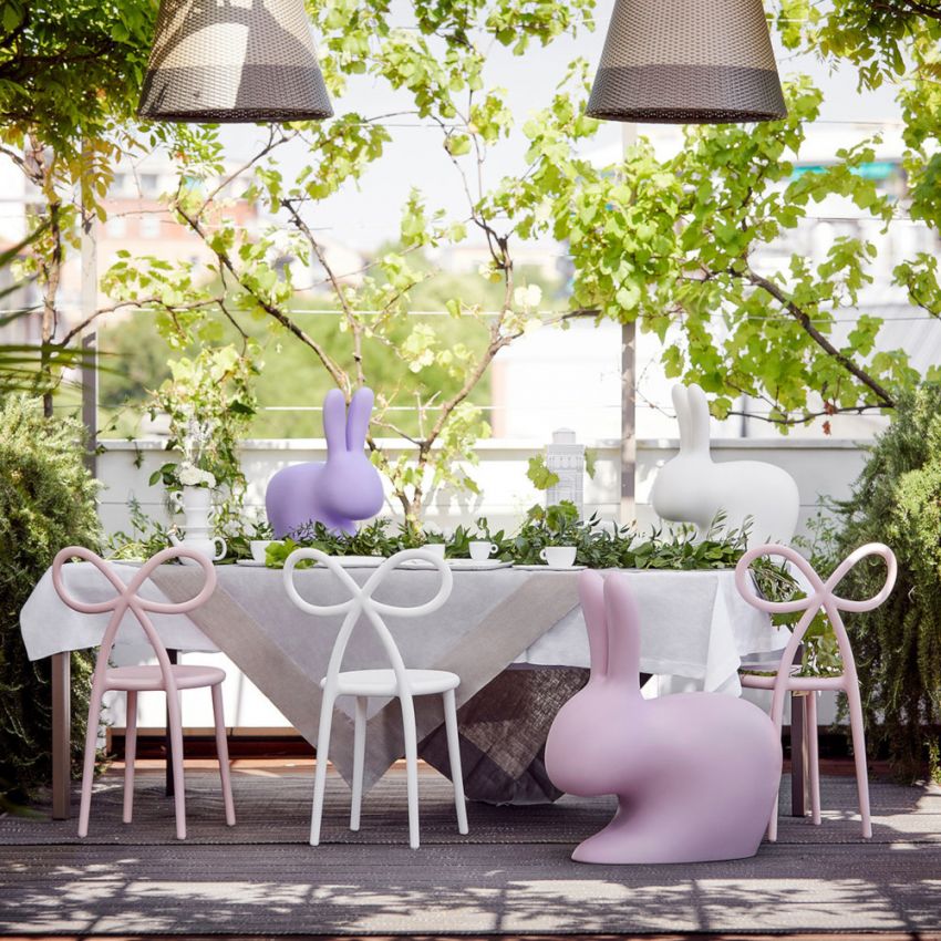 A styled outdoor dining scene with a table covered in a white cloth, four bow-back Qeeboo Ribbon chairs in pink and white, a pastel pink bunny-shaped chair, and decorative bunny sculptures in purple and white beneath hanging lamps and leafy trees.