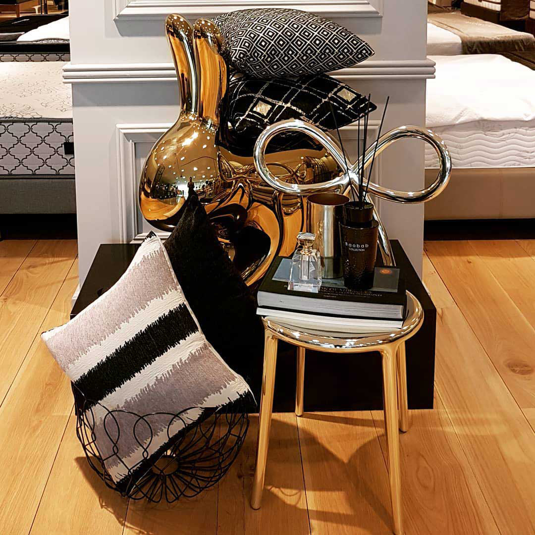 A styled interior scene with a gold-toned side table holding books, a black bottle, and a black metal diffuser, alongside a gold rabbit figurine and patterned throw pillows on a black pedestal in a bedroom setting. Ribbon silver chair by Qeeboo.