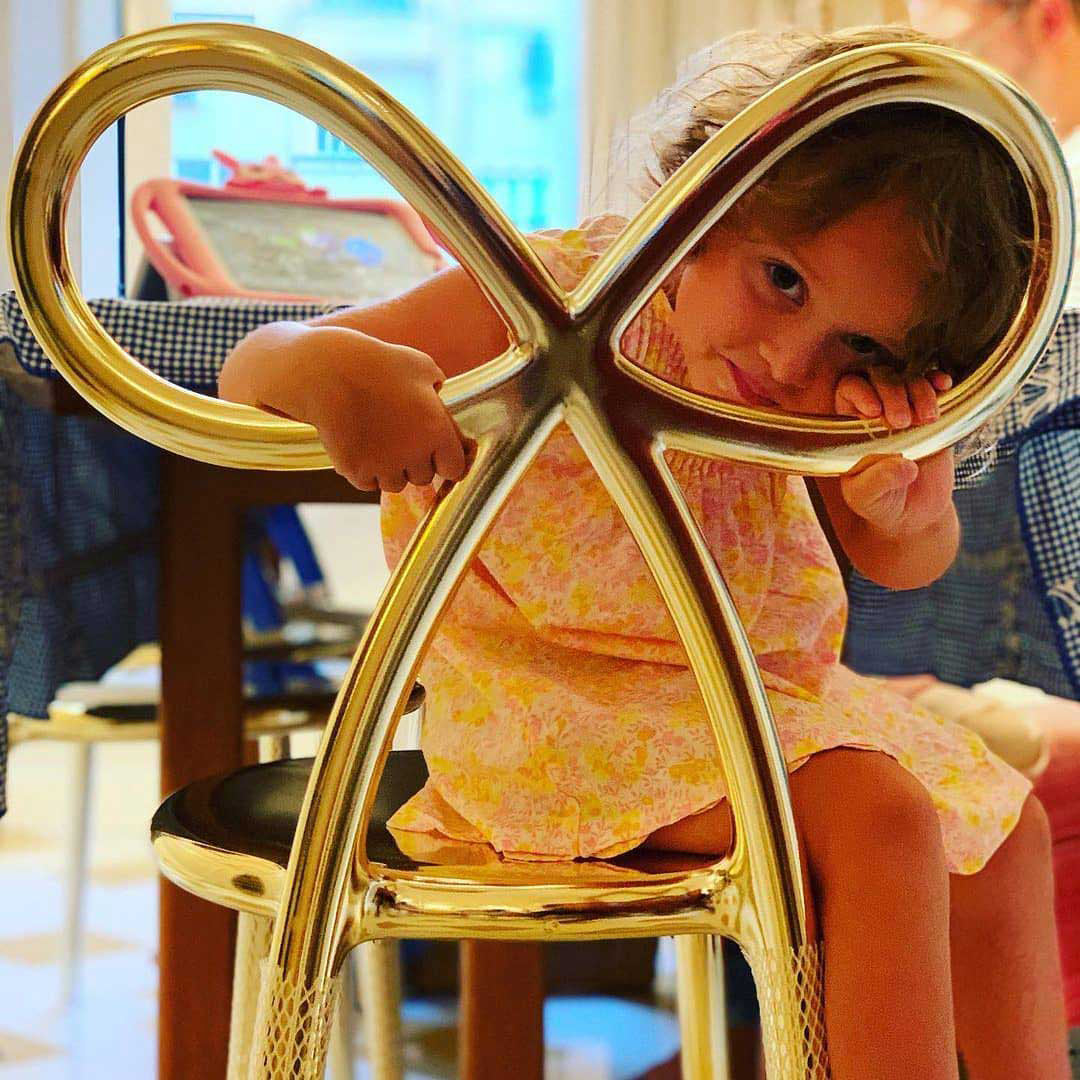 A young child in a pink floral dress sits on a highly polished gold Ribbon metal chair with an abstract looped backrest. Qeeboo Made in Italy