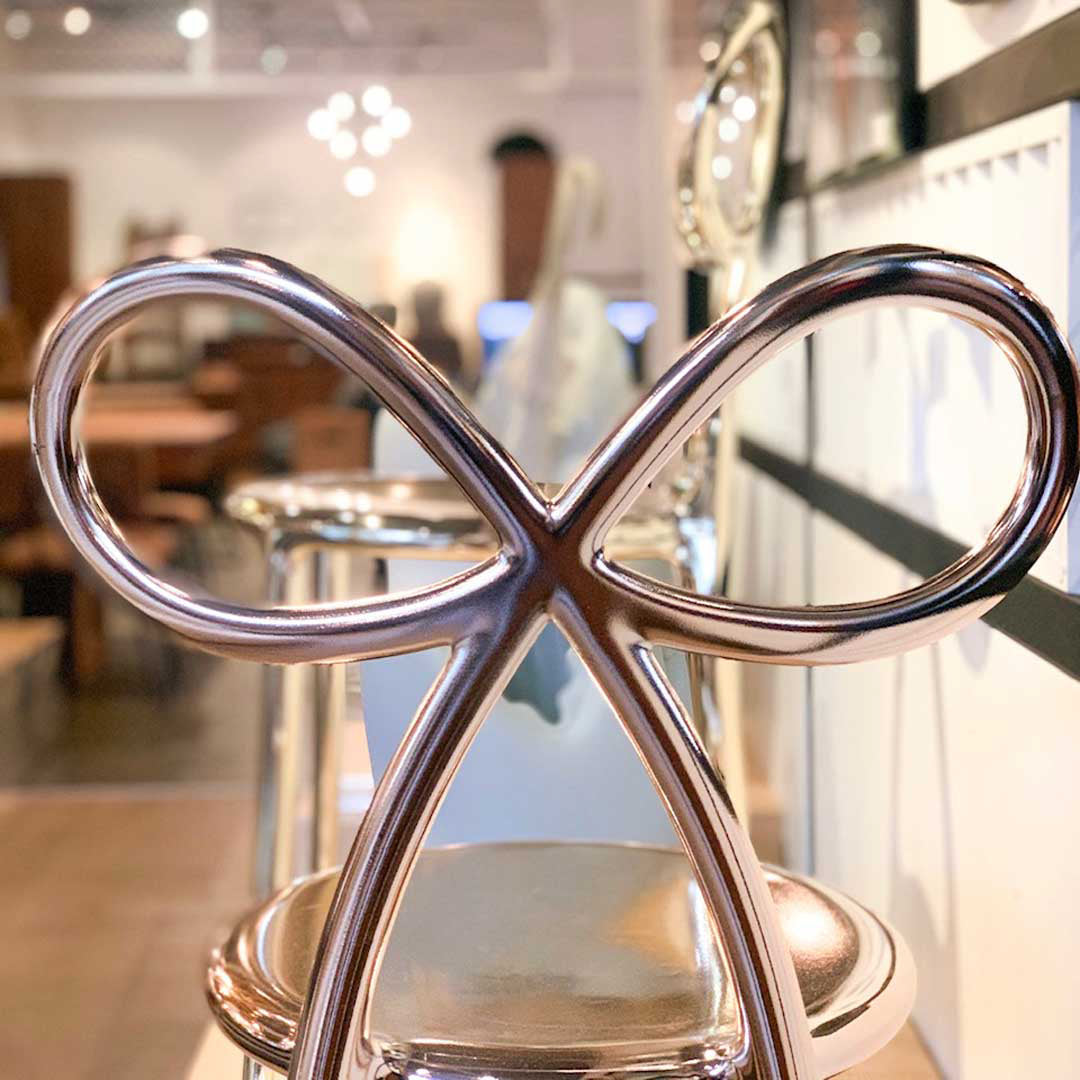 A modern pink gold metal chair with a sculptural infinity symbol backrest, shown in a softly blurred retail interior. Ribbon by Qeeboo.