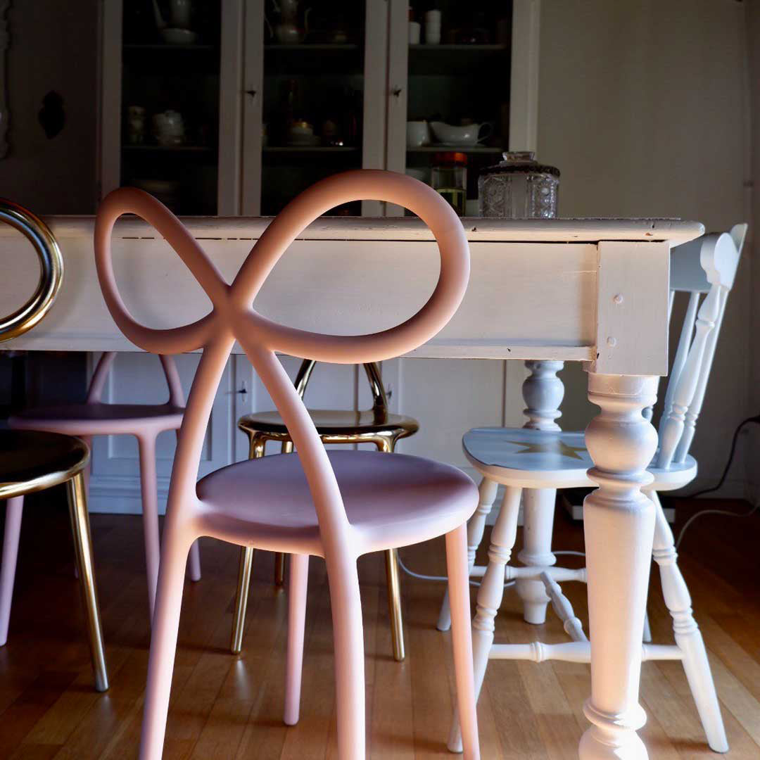 A pink chair with a looped back positioned in front of a white wooden dining table in a home interior setting. Ribbon chair by QEEBOO.